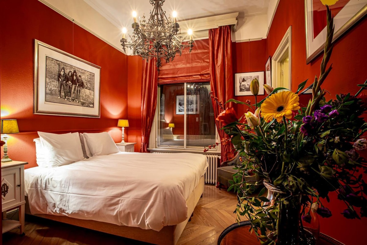 A hotel room with a large bed, a chandelier, a window with curtains, and a floral arrangement.