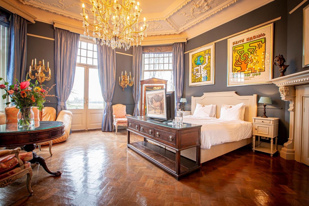A luxurious bedroom with a large bed, a chandelier, a fireplace, and artwork on the walls.