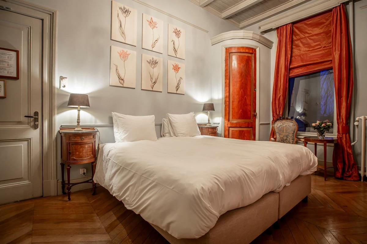 A bedroom with a large bed, two bedside tables, a nightstand, a wooden wardrobe, a window with red curtains, and wall-mounted artwork.