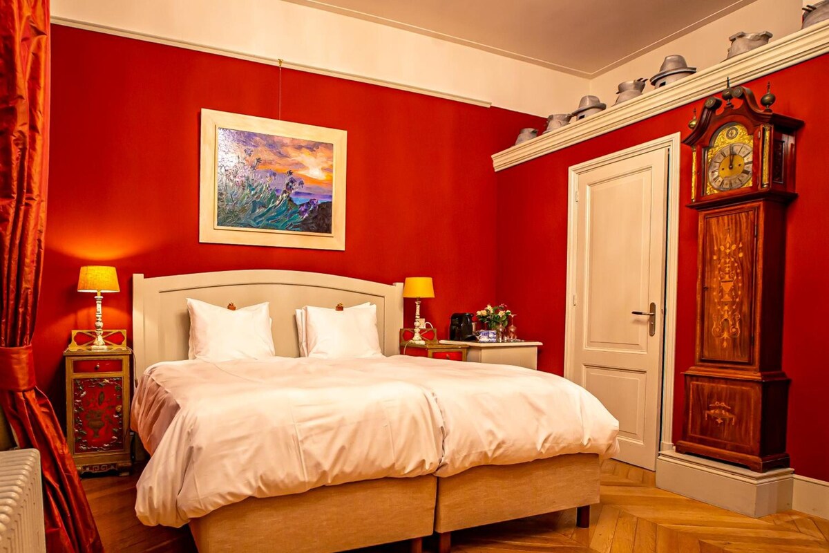 Un dormitorio con una cama grande, dos mesitas de noche con lámparas, un cuadro en la pared, una puerta y un reloj de madera alto.