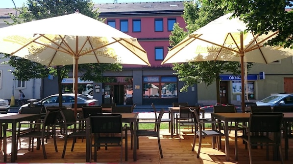 An outdoor seating area with tables and chairs under umbrellas, a building with a sign 'SQUASH FITNESS' in the background, and a parked car.