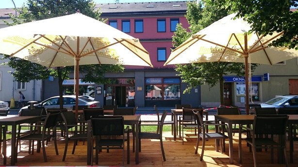 An outdoor seating area with tables and chairs under umbrellas, a building with a sign 'SQUASH FITNESS' in the background, and a parked car. - Hotel Kaskada (Ledeč nad Sázavou)