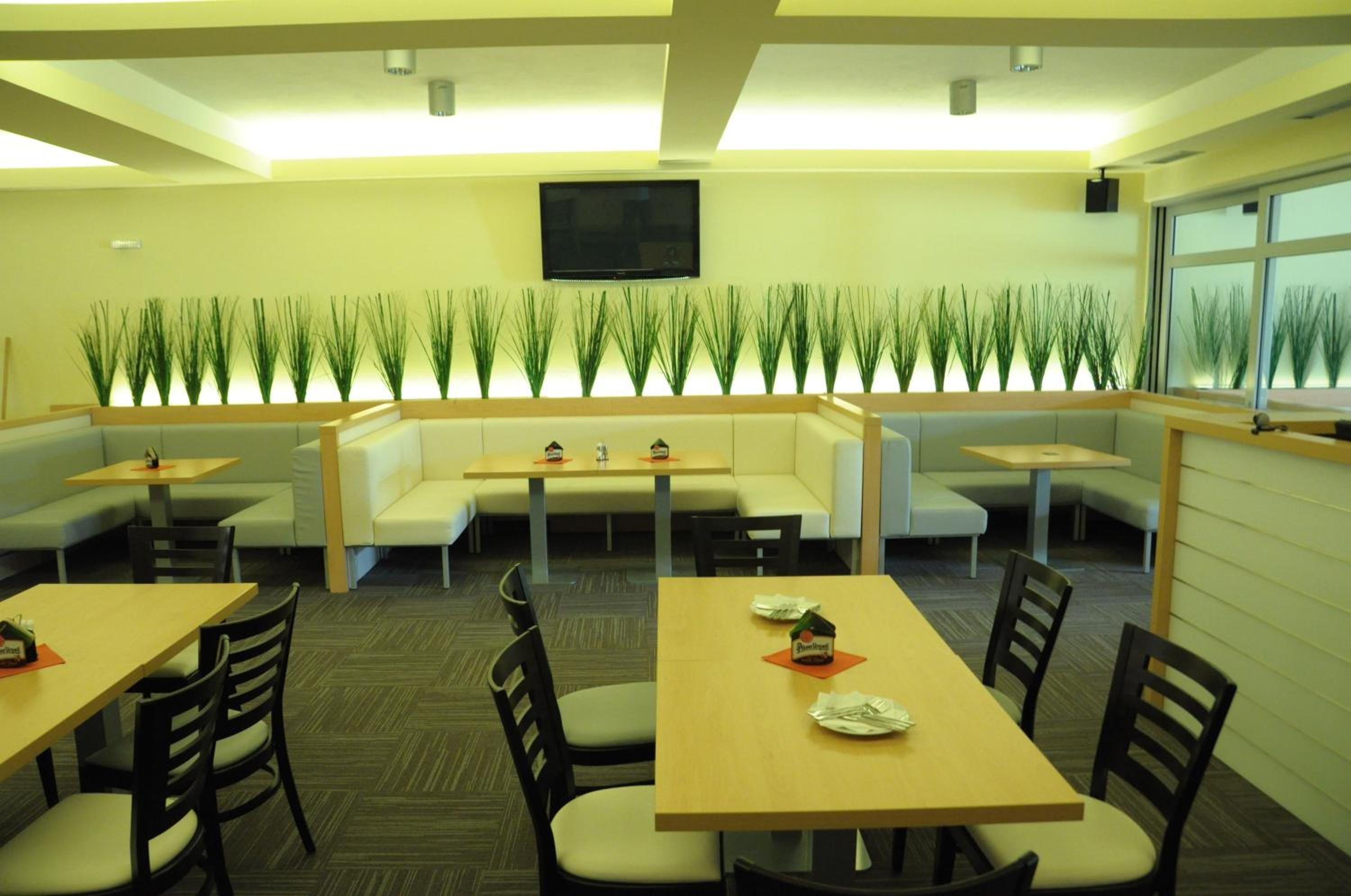 A restaurant with booths and tables, a flat-screen TV mounted on the wall, and a row of green plants along the back wall.
