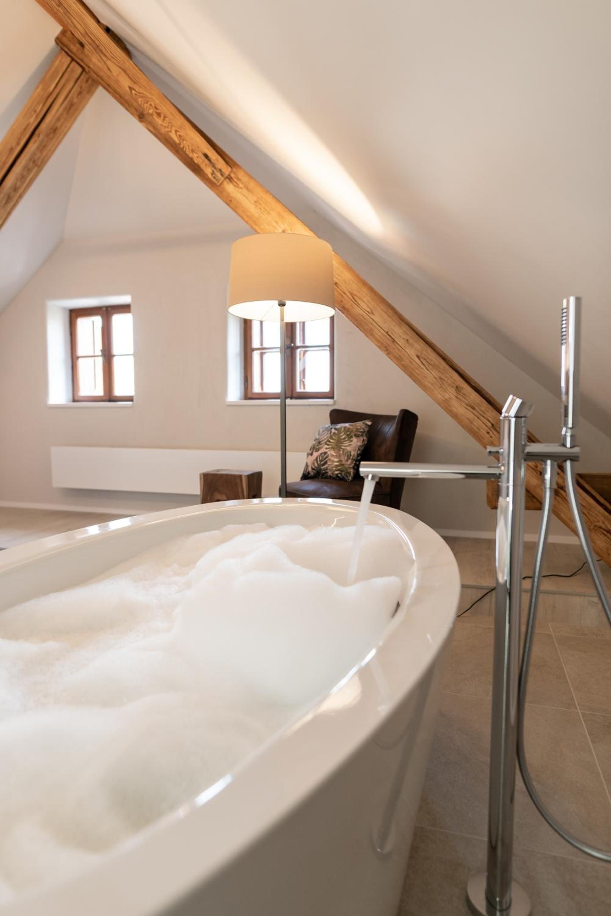 Ein Zimmer mit einer großen Badewanne voller Wasser, einer Holzbalkendecke und einem Sessel mit Kissen.