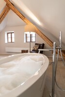 Una habitación con una bañera grande llena de agua, techo de madera y una silla con un cojín.