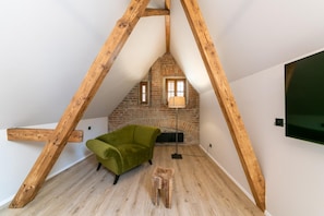Un dormitorio tipo loft con vigas de madera a la vista, una pared de ladrillo, un sillón verde, un taburete de madera y una lámpara de pie.