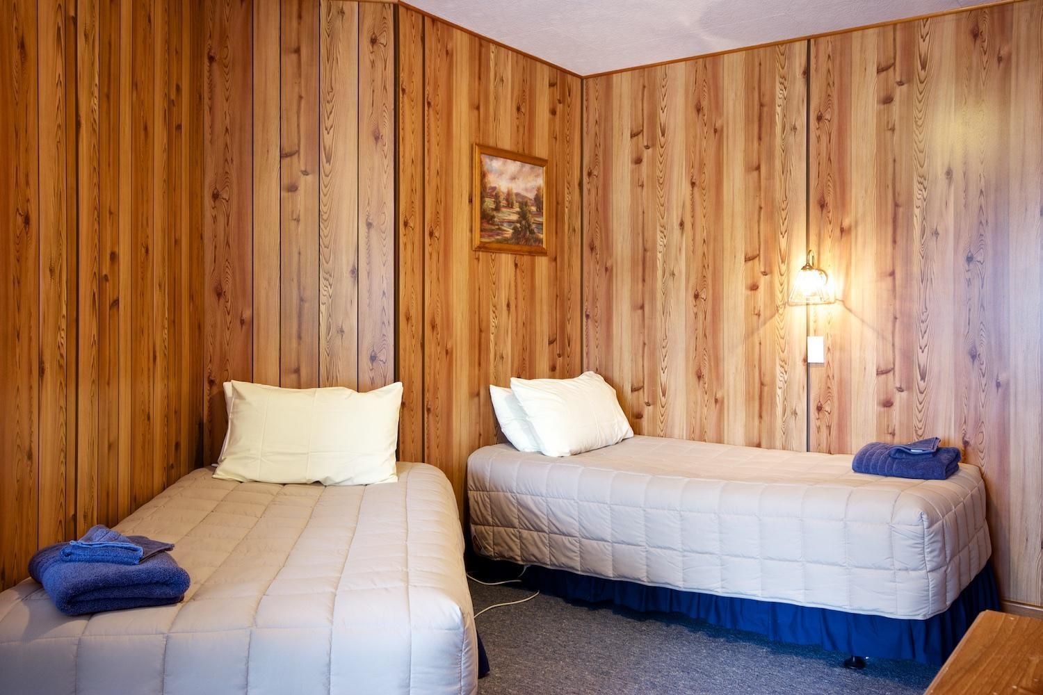 A room with two beds, wooden walls, a painting, and a wall-mounted light fixture.