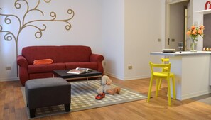 Una sala de estar con un sofá rojo, un reposapiés negro, una alfombra a cuadros, un sillón amarillo y un peluche en la alfombra.
