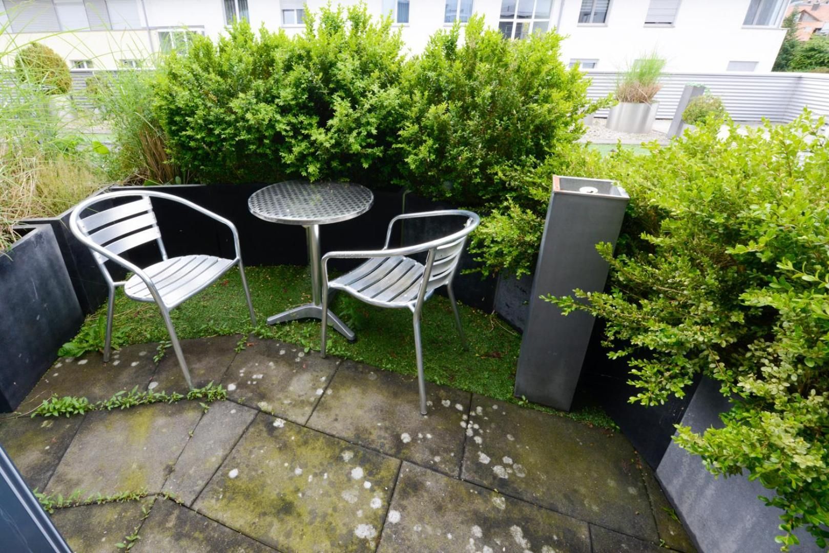 A small outdoor seating area with two metal chairs and a round table, surrounded by greenery and a stone floor.