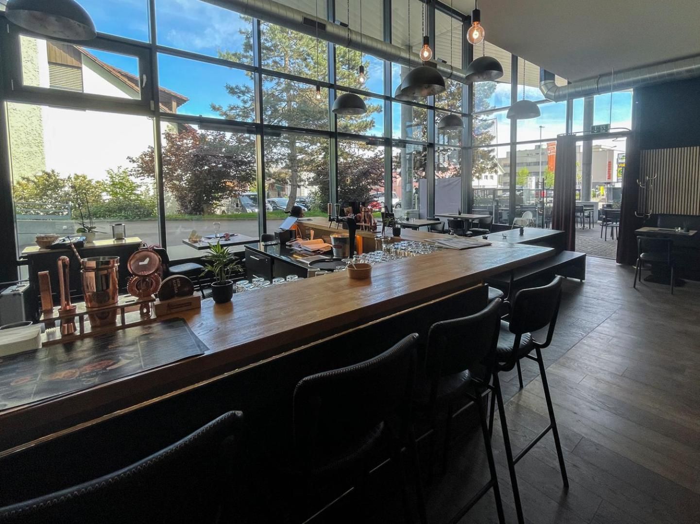 A modern bar area with a long counter, multiple bar stools, and large windows offering a view of the street outside.
