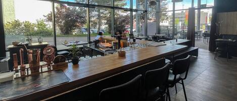 A modern bar area with a long counter, multiple bar stools, and large windows offering a view of the street outside.