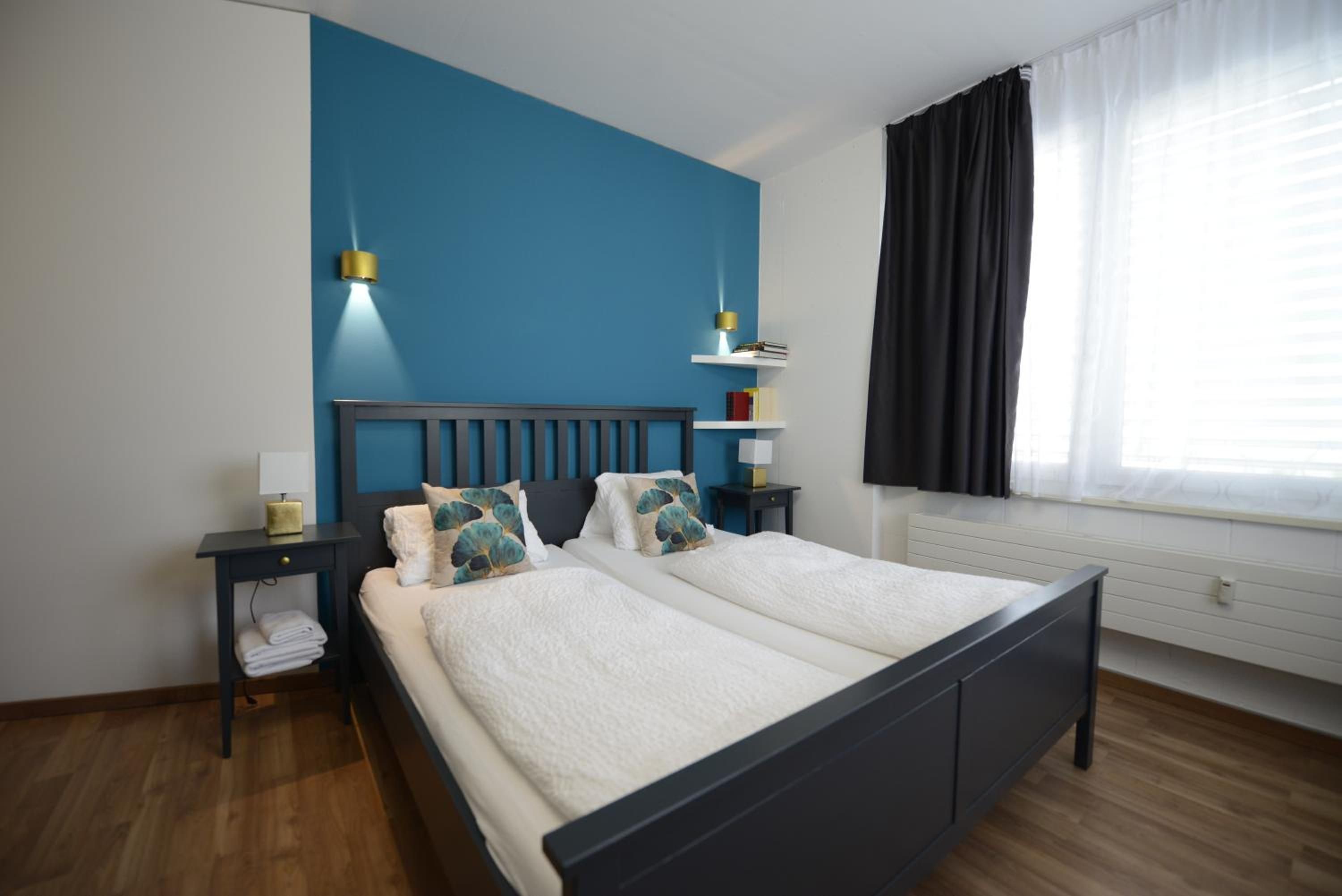 A double bed with a dark headboard, two bedside tables with lamps, a window with curtains, and a blue accent wall.