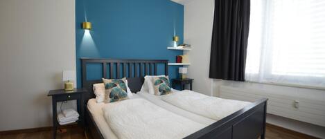A double bed with a dark headboard, two bedside tables with lamps, a window with curtains, and a blue accent wall.