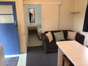 Espaço de estar compacto com sofá, mesa pequena, cama com manta listrada e porta que dá para outro cômodo.