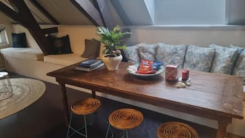 Een knus loft-achtig vertrek met een houten eettafel, drie rieten barstoelen, een potplant, snacks en een raam met uitzicht.