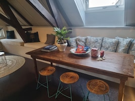 Ein gemütliches Loft-Zimmer mit einem Holzesstisch, drei Korbstühlen, einer Zimmerpflanze, Snacks und einem Fenster mit Blick.