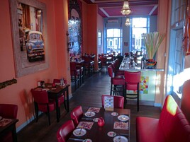Ein Restaurant mit roten Stühlen und Tischen, einer Bar und großen Fenstern, die viel Tageslicht hereinlassen.