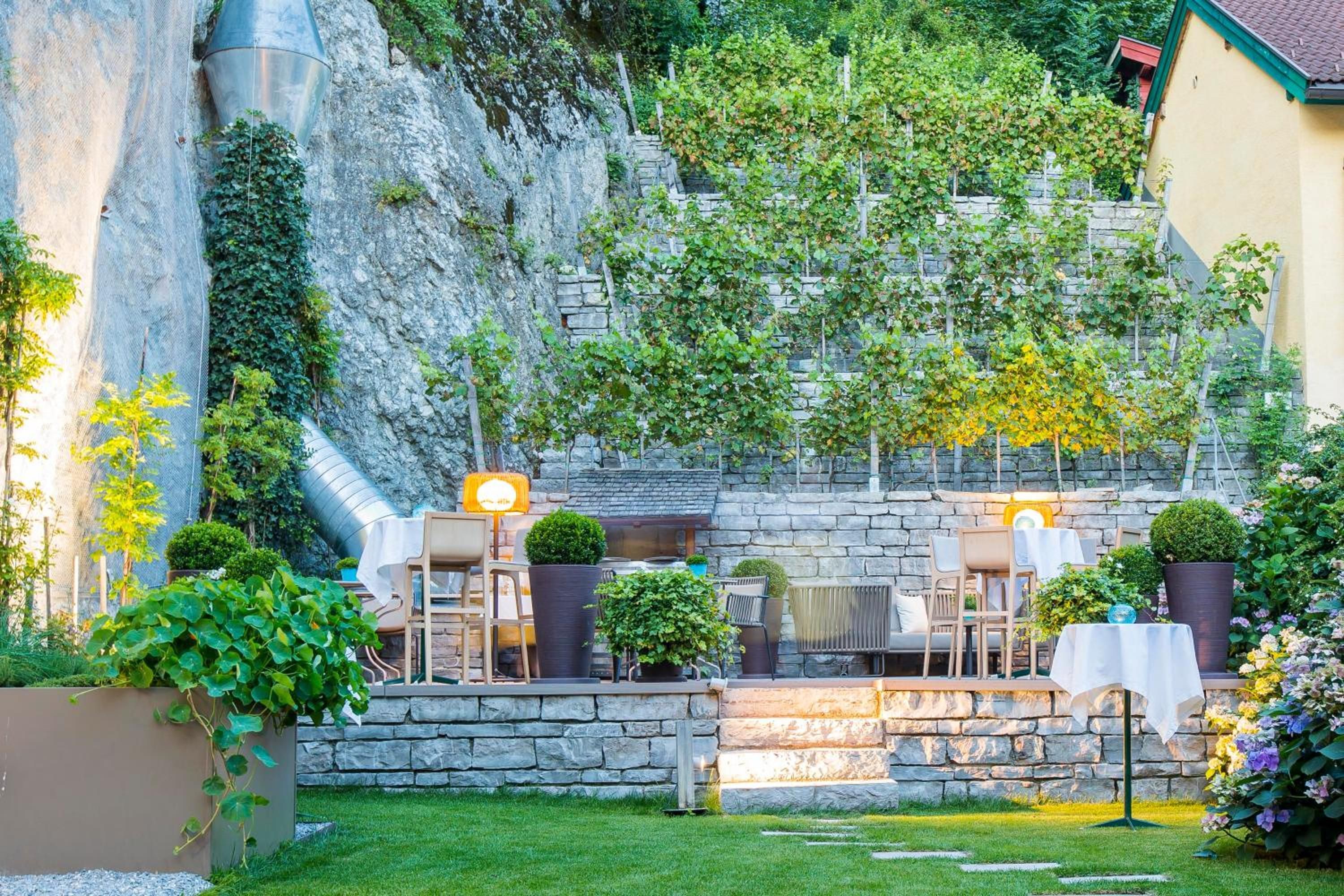 A stone wall with climbing plants, outdoor seating with tables and chairs, and a small garden area.