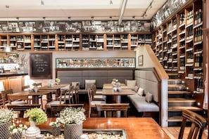 Un bar con estantes de madera llenos de botellas, una zona de estar con mesas y sillas, y una pizarra con texto.
