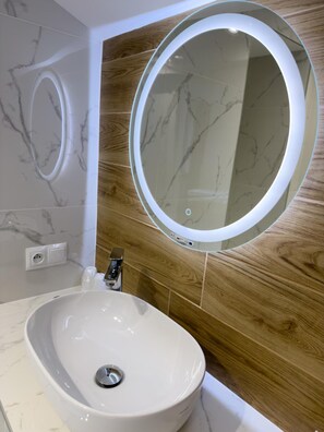 A bathroom with a white marble sink, a wooden panel wall, and a round mirror with a light. - Hotel Sjesta (Mikołów)
