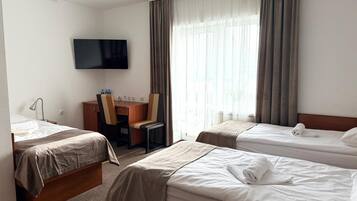 A hotel room with two single beds, a desk with a chair, a flat-screen TV, and a large window with curtains.