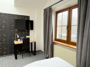 A hotel room with a bed, a desk with a chair, a flat-screen TV mounted on the wall, and a window with curtains. - Hotel Sjesta (Mikołów)