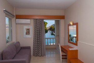 A room with a sofa, a desk, a chair, a window with a view of the sea, and a balcony.