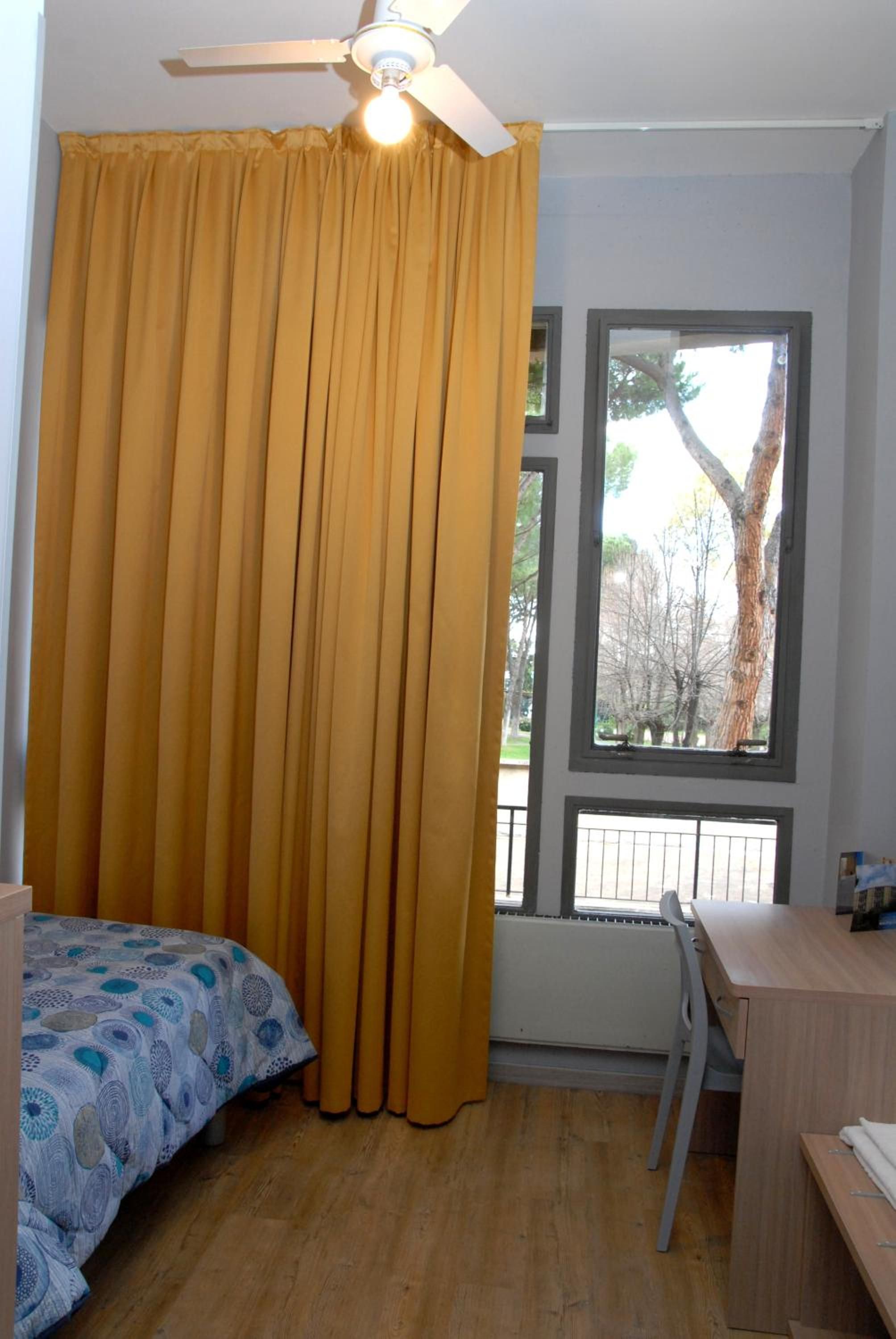 A bedroom with a bed, a desk, a chair, a window with a view of trees, and a ceiling fan.