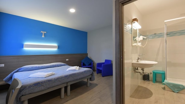 A hotel room with a bed, two chairs, a bathroom with a sink and mirror, and a blue wall.