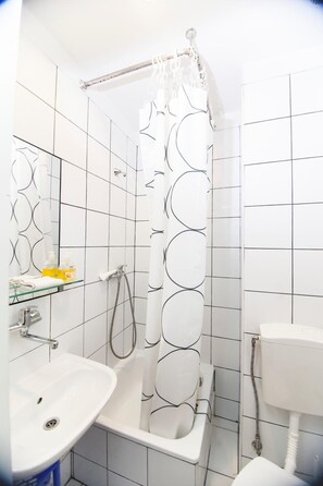 A bathroom with white tiled walls, a shower with a circular pattern curtain, a glass shelf with toiletries, and a white sink. - Akacja Pokoje Go Ocinne (Kraków)