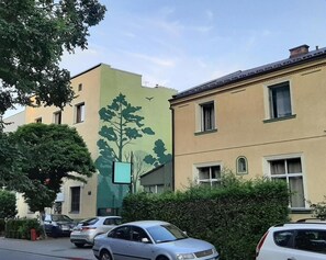 A street view with parked cars, a building with a tree mural, and a hedge. - Akacja Pokoje Go Ocinne (Kraków)