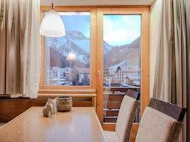 A room with a wooden table, chairs, a lamp, and a window with a view of mountains and buildings.