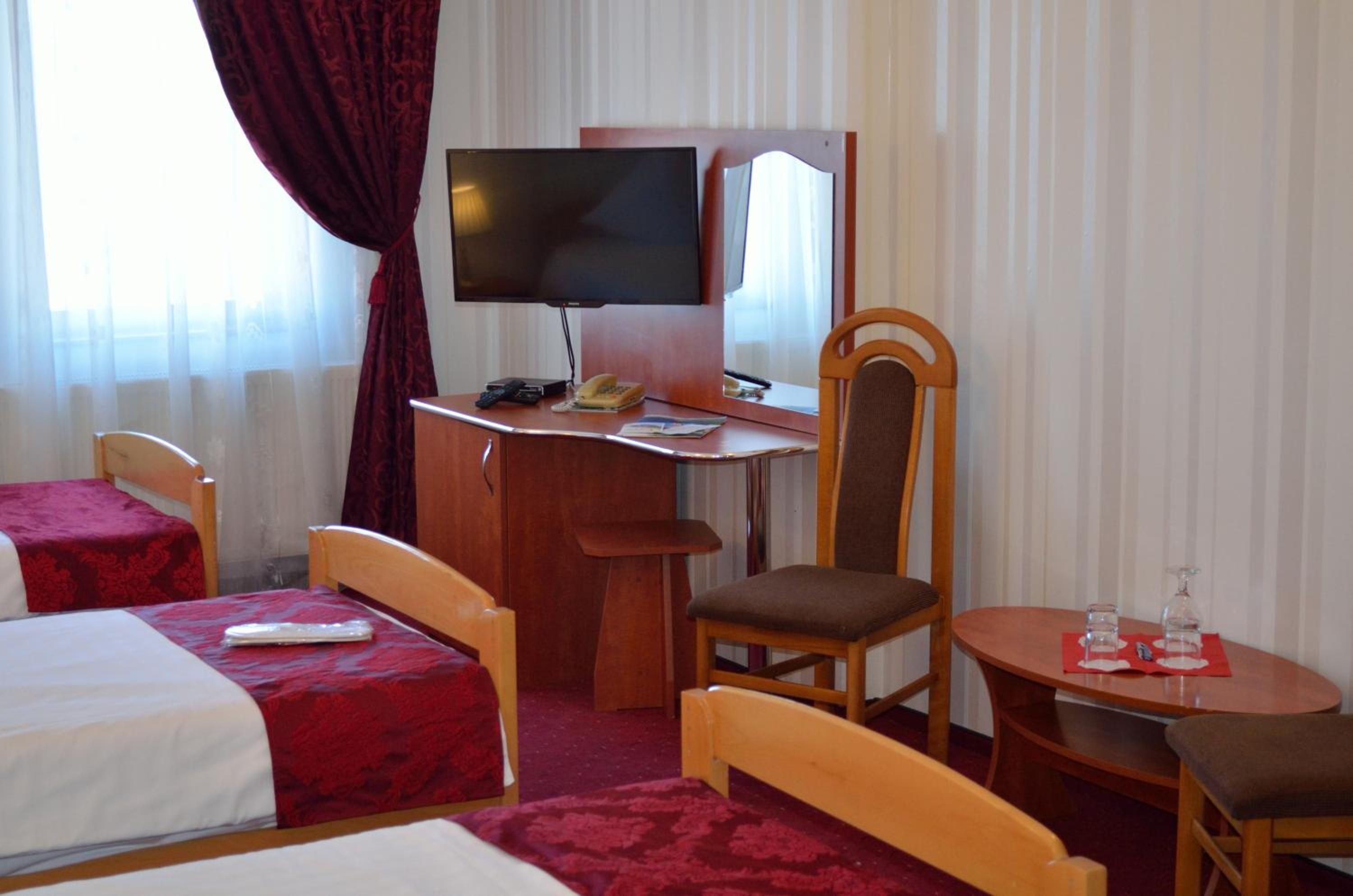 A hotel room with a bed, a desk with a television, a chair, a small table with a glass, and a window with curtains.