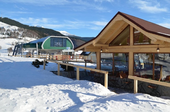 A wooden cabin with a glass enclosure, a covered outdoor seating area, and a green building in the background.