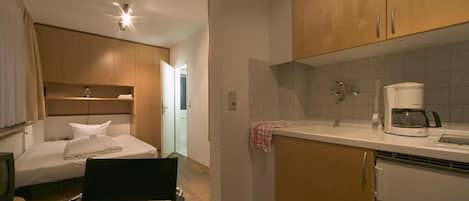 A compact hotel room with a bed, a small desk, a chair, a kitchenette with a coffee maker, and wooden cabinets.