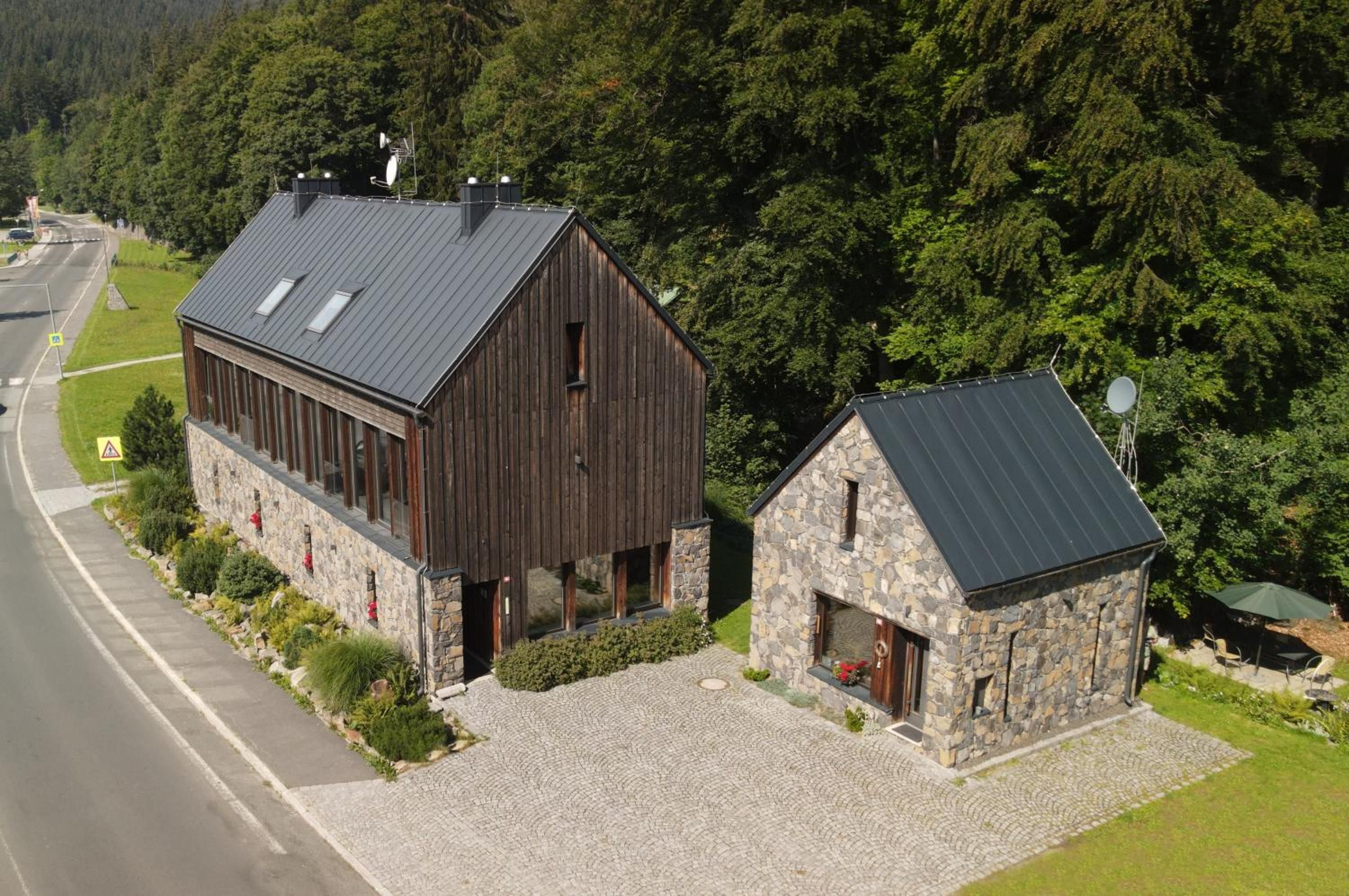 A two-story wooden building with a stone annex, surrounded by a cobblestone driveway and lush greenery.
