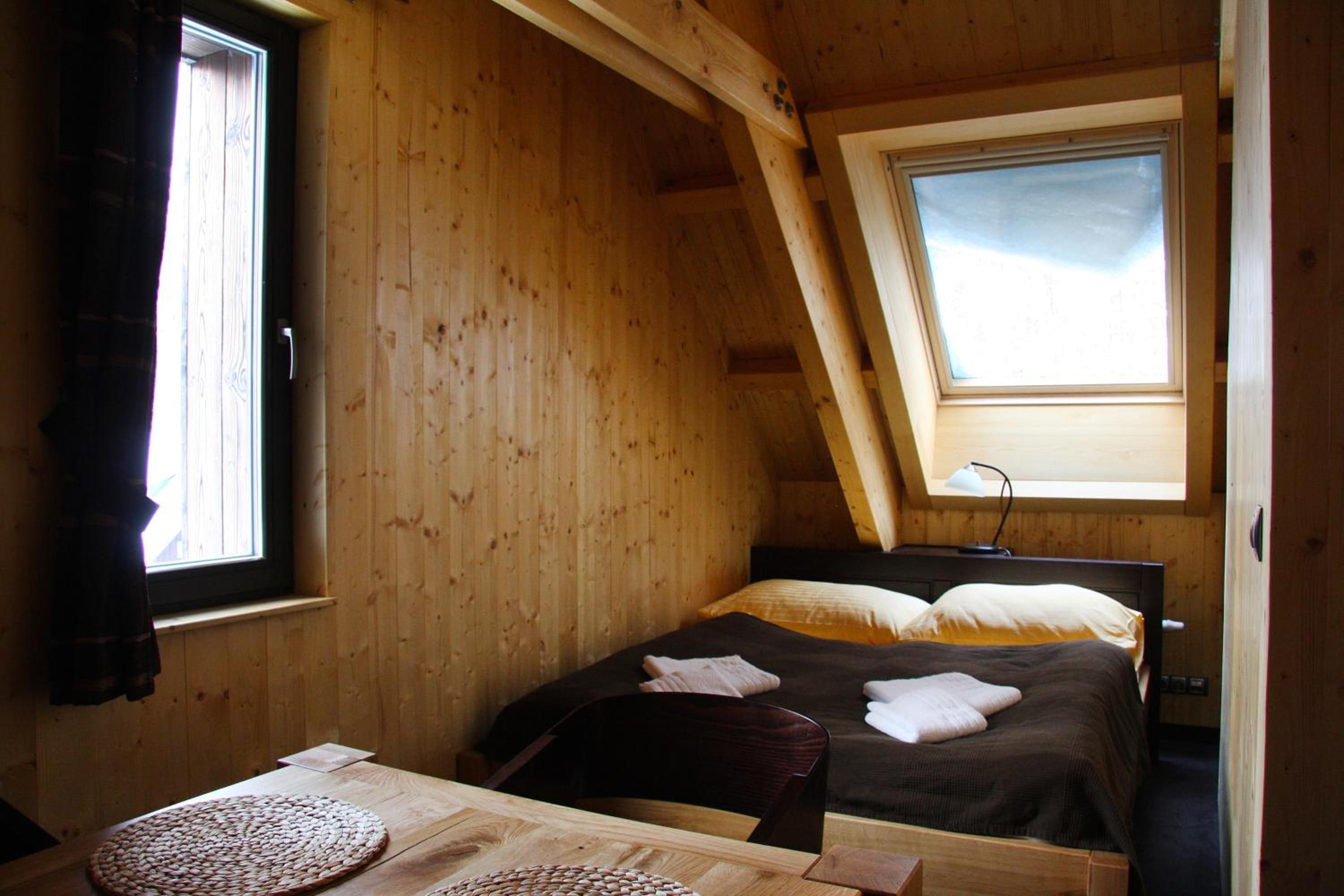A wooden room with a sloped ceiling, a bed with pillows, a table with a woven placemat, and a window with curtains.