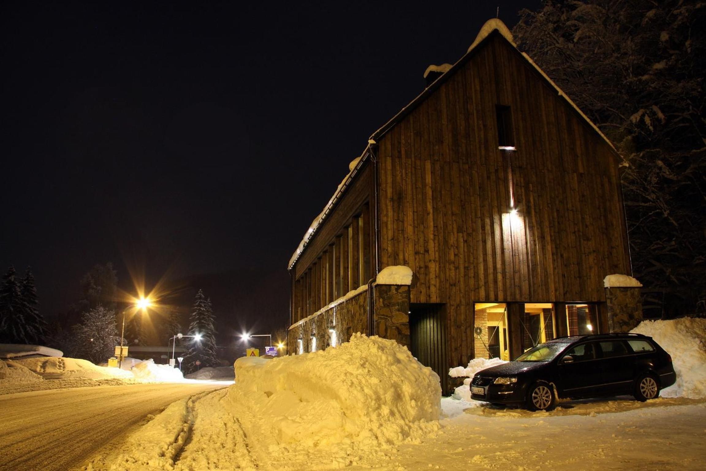 Un edificio de madera con el suelo cubierto de nieve, un coche aparcado delante y un montículo de nieve a un lado.