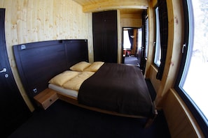 Una cama individual con cabecera oscura y dos almohadas, suelo de madera y ventana con cortinas.