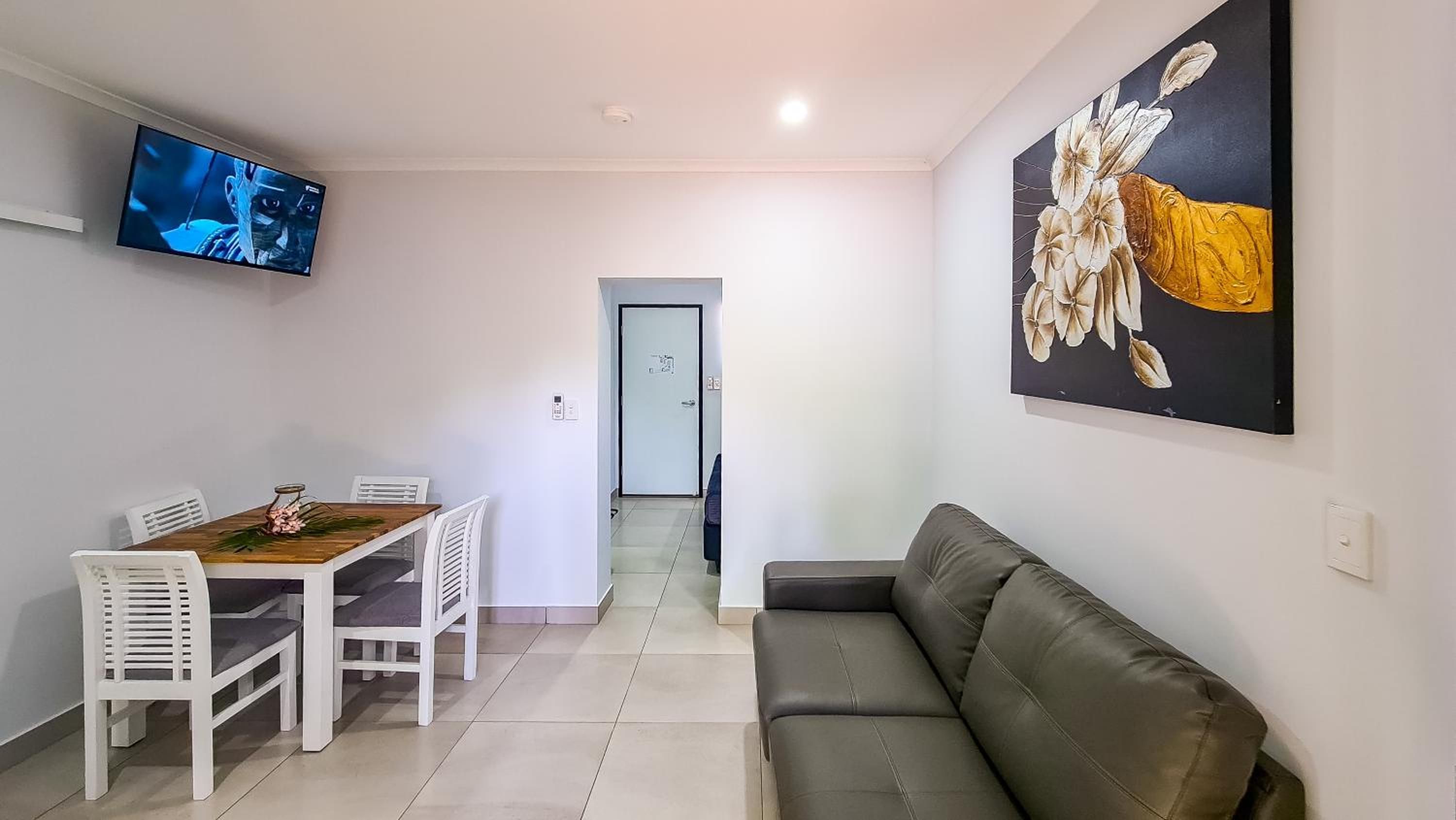 A living room with a sofa, a dining table with chairs, a TV mounted on the wall, and a door leading to another room.