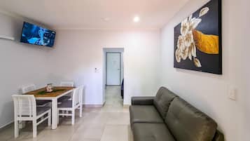 A living room with a sofa, a dining table with chairs, a TV mounted on the wall, and a door leading to another room.