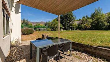 An outdoor patio with a table and chairs, a shade sail, and a view of a grassy field and trees.
