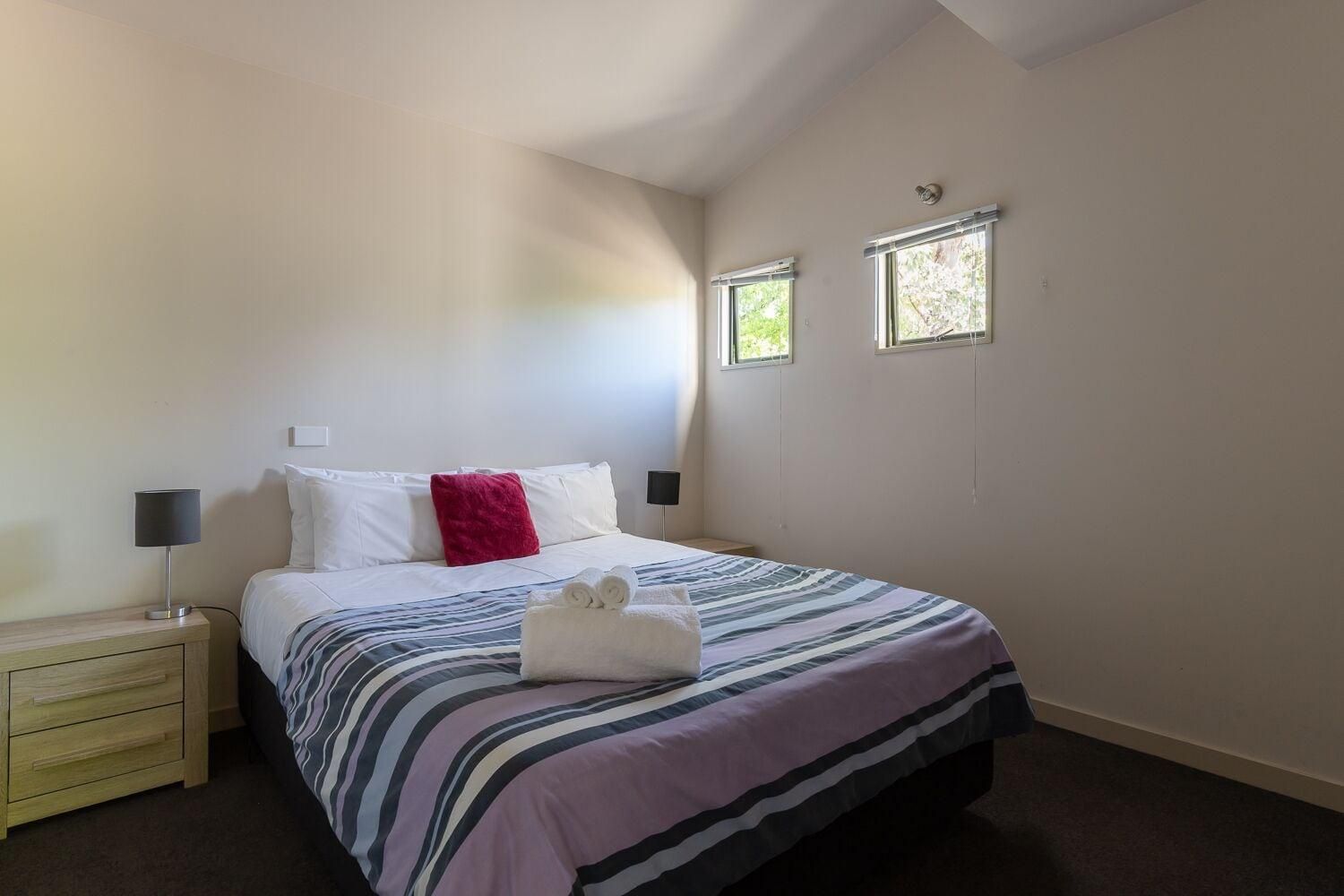 Una habitación de hotel con una cama, lámparas de noche, una mesita de noche y dos ventanas.