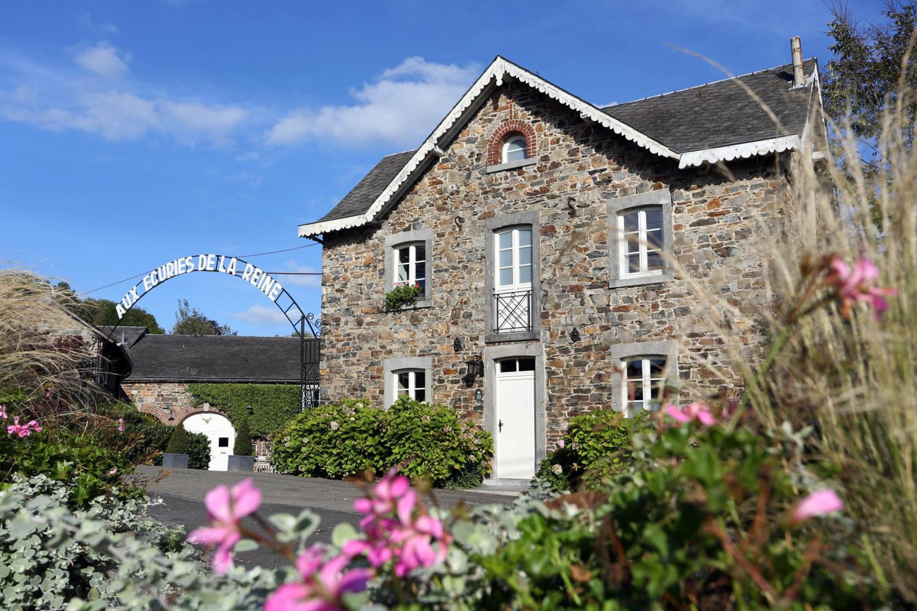 Un bâtiment en pierre avec une porte blanche et de nombreuses fenêtres, entouré de verdure et sous un ciel dégagé.