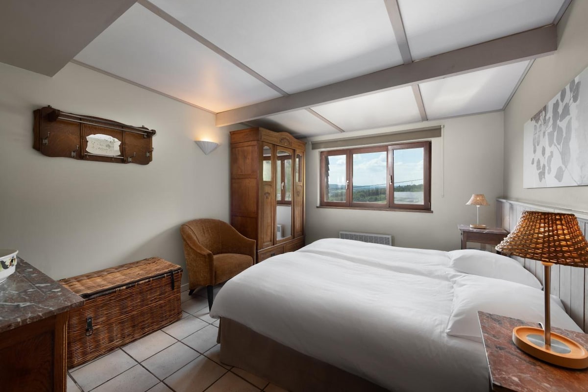 A bedroom with a bed, a chair, a wardrobe, a desk with a lamp, and a window with a view.