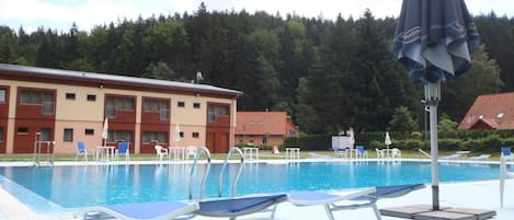 Una piscina con sillones de descanso y sombrillas, rodeada de un edificio y árboles.
