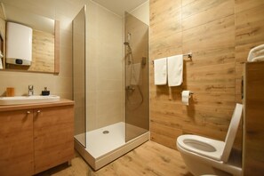 A modern bathroom with a glass-enclosed shower, wooden wall paneling, a white toilet, and a wooden vanity with a sink. - Korzo Apartmani (Podgorica)