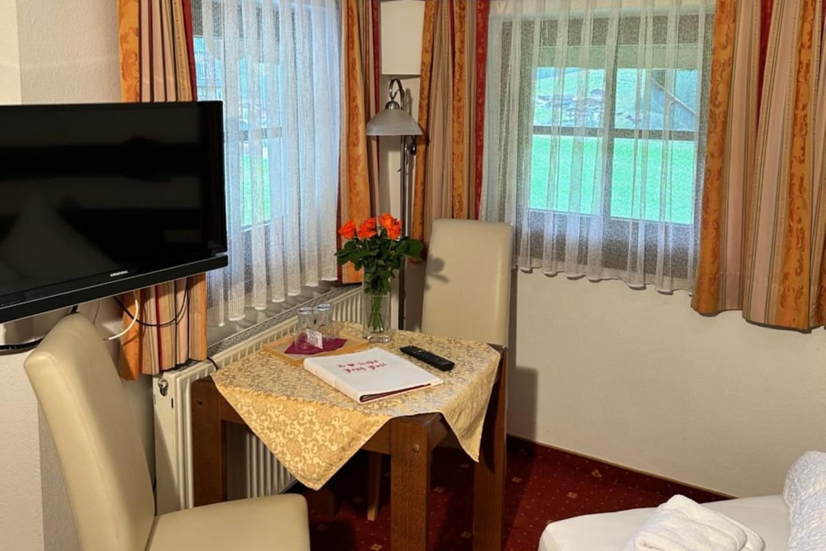 Ett hotellrum med en säng, ett skrivbord med en stol, en tv, ett bord med en vas med blommor och gardiner.
