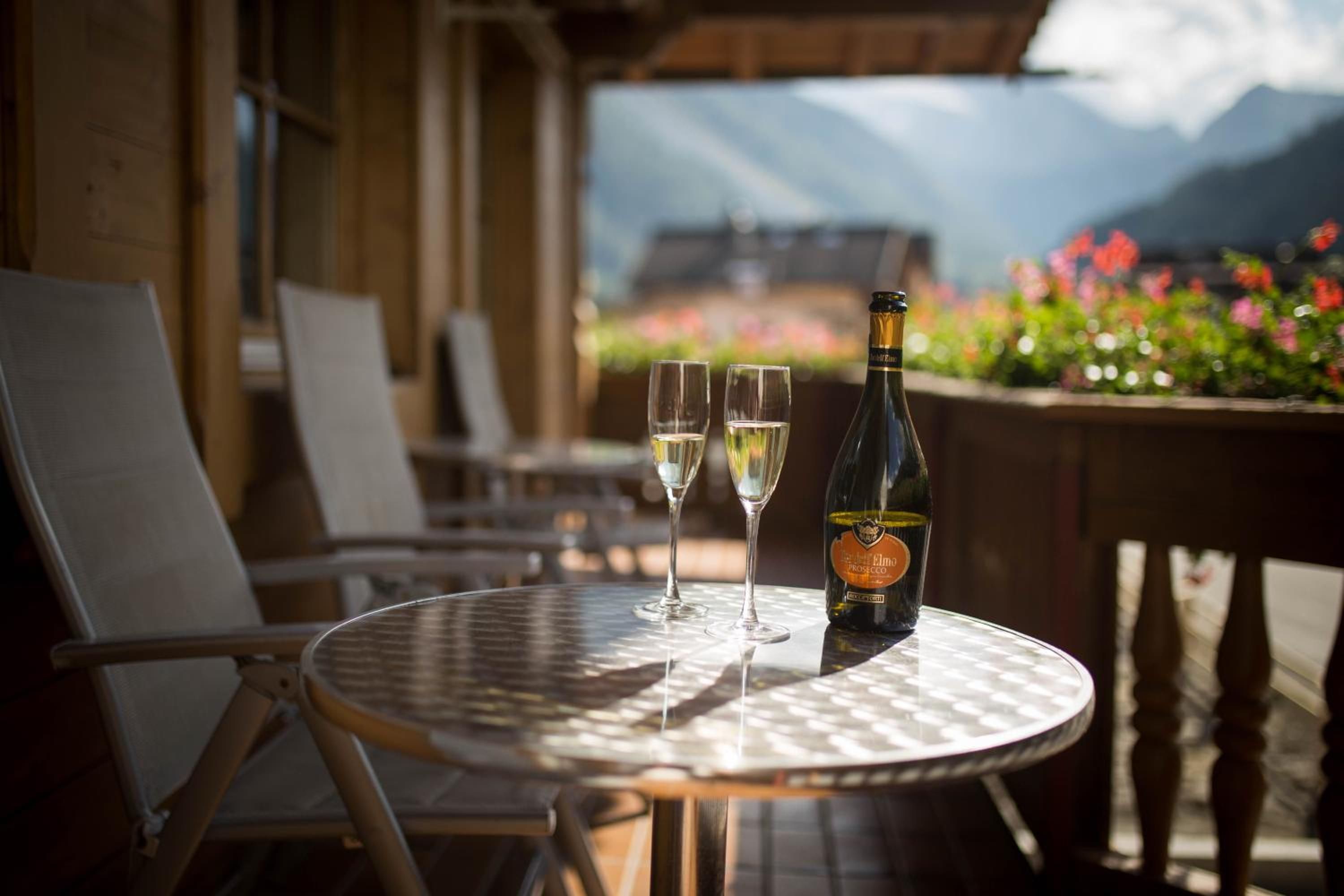 A table with a bottle and two glasses on a round table, set in a room with chairs and a view of mountains and flowers.