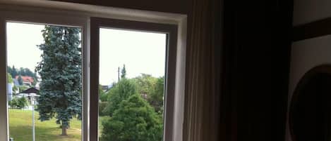 Habitación de hotel con cama, escritorio, lámpara y vista a la calle y zonas verdes a través de una puerta corredera.